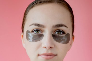 Baş portresi. Göz altı nemlendirici maskeli gülümseyen genç bir kadın. Kameraya bakıp cilt bakımının keyfini çıkarıyor..