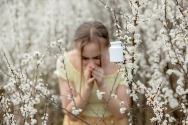 Genç bir kadın, çiçek açan ağacın önünde burun sümkürerek ve mendile hapşırarak. İnsanları etkileyen mevsimsel alerjik maddeler. Güzel bayanın rhinitis 'i var..