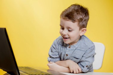 Mutlu çocuk dizüstü bilgisayarla masada oturuyor ve sarı arka planda okula hazırlanıyor. Çevrimiçi öğrenme. Çevrimiçi okul