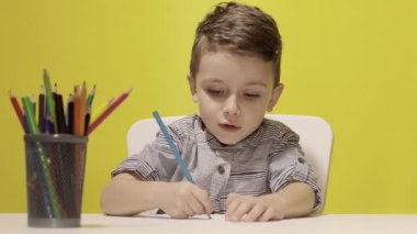 Masada gülümseyen küçük çocuk sarı arka planda annesi için pastel boyalarla resim çiziyor. Yaratıcılık konsepti. Okula geri dön.