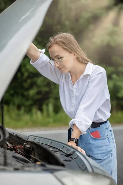 Young woman standing near broken down car with popped up hood having trouble with her vehicle. Waiting for help tow truck or technical support. A woman calls the service center.