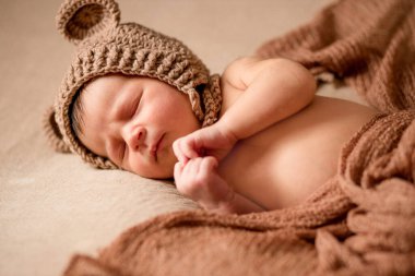 Yeni doğan, örgü şapkalı uyuyan bebek kahverengi kumaşın üzerinde yatıyor..