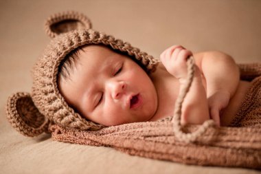 Yeni doğan, örgü şapkalı uyuyan bebek kahverengi kumaşın üzerinde yatıyor..