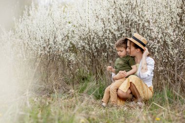 Mutlu anne ve oğul birlikte eğleniyor. Anne oğluna nazikçe sarılıyor. Arka planda beyaz çiçekler açar. Anneler Günü..