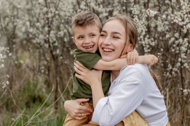 Mutlu anne ve oğul birlikte eğleniyor. Anne oğluna nazikçe sarılıyor. Arka planda beyaz çiçekler açar. Anneler Günü..