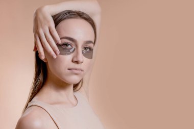 Bej arka planda gözlerinin altında uygulamalı siyah güzellik bantlarıyla poz veren genç kadın portresi.