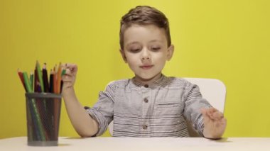 Masada gülümseyen küçük çocuk sarı arka planda annesi için pastel boyalarla resim çiziyor. Yaratıcılık konsepti. Okula geri dön.