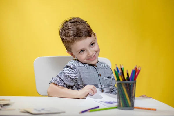 Smiling little boy at the table draw with crayons for mum on yellow background. Creativity concept. Back to school