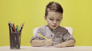 Masada gülümseyen küçük çocuk sarı arka planda annesi için pastel boyalarla resim çiziyor. Yaratıcılık konsepti. Okula geri dön.