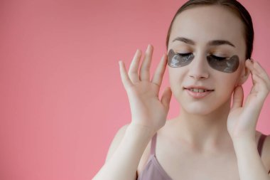 Baş portresi. Göz altı nemlendirici maskeli gülümseyen genç bir kadın. Kameraya bakıp cilt bakımının keyfini çıkarıyor..