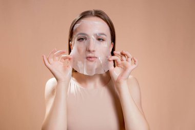 Yüzünde kahverengi bir arka planda kozmetik maske olan genç bir kadın. Deri durumunu iyileştirmek için yüz maskesi. Yüz bakımı. Kozmetik gevşeme.