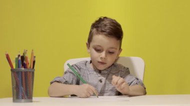 Masada gülümseyen küçük çocuk sarı arka planda annesi için pastel boyalarla resim çiziyor. Yaratıcılık konsepti. Okula geri dön.