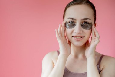 Baş portresi. Göz altı nemlendirici maskeli gülümseyen genç bir kadın. Kameraya bakıp cilt bakımının keyfini çıkarıyor..