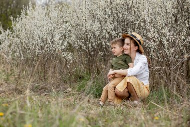 Mutlu anne ve oğul birlikte eğleniyor. Anne oğluna nazikçe sarılıyor. Arka planda beyaz çiçekler açar. Anneler Günü..