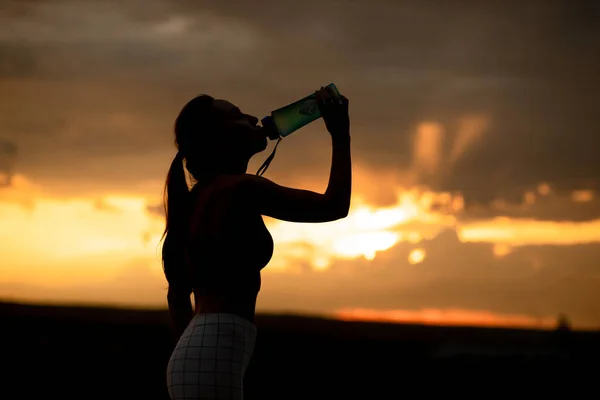 Silhouette of girl in sportswear drinks water from a bottle at sunset — Stock Photo, Image