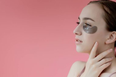 Baş portresi. Göz altı nemlendirici maskeli gülümseyen genç bir kadın. Kameraya bakıp cilt bakımının keyfini çıkarıyor..