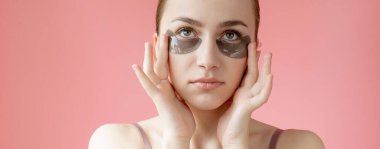 Baş portresi. Göz altı nemlendirici maskeli gülümseyen genç bir kadın. Kameraya bakıp cilt bakımının keyfini çıkarıyor..