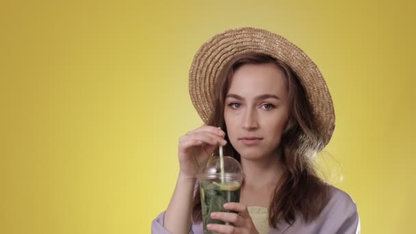 黄色のスタジオを背景にプラスチックカップからモヒートカクテルを飲む驚きの女性 — ストック動画