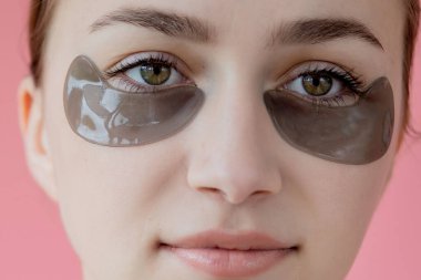 Baş portresi. Göz altı nemlendirici maskeli gülümseyen genç bir kadın. Kameraya bakıp cilt bakımının keyfini çıkarıyor..