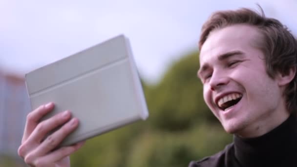 Junger Mann mit Tablet-Computer im Park sitzend, plaudernd, surfend, studierend. Glücklich aussehen. — Stockvideo