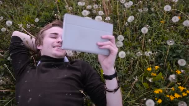 Ung glad student med hjälp av Tablet PC i parken, liggande i gräset i maskrosor, ler och skrattar. — Stockvideo