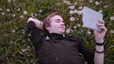 Gülümseyen ve gülerek dandelions, çimlere yatan genç mutlu öğrenci park, tablet kullanma.
