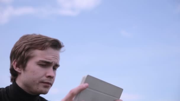 Jeune homme avec tablette assis dans le parc, bavardant, surfant, claquant. Ciel sur le terrain de pêche . — Video