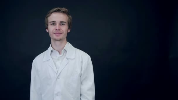 Smiling Doctor showing thumbs up, standing on a black background putting on a stethoscope. Packshot. — Stock Video