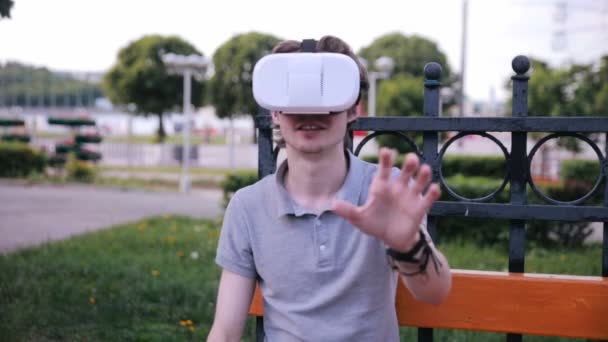 Young handsome man wearing virtual reality goggles sitting in a city park. — Stock Video