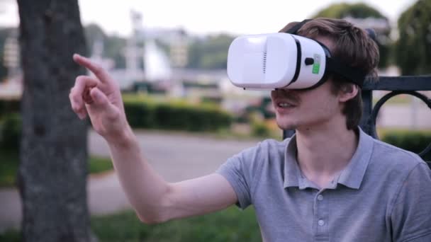 Young handsome man wearing virtual reality goggles sitting in a city park. — Stock Video