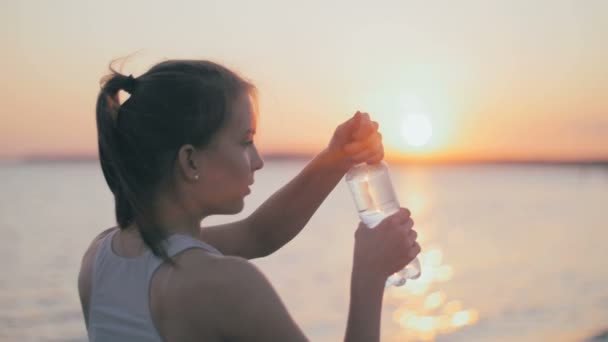 Chica atleta joven bebiendo agua después de correr fitness, entrenamiento de jogger al atardecer de verano. Movimiento lento . — Vídeo de stock