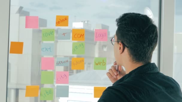 Joven hombre guapo escribiendo en notas adhesivas en la ventana panorámica en la oficina moderna. Lluvia de ideas, nombrar . — Vídeos de Stock