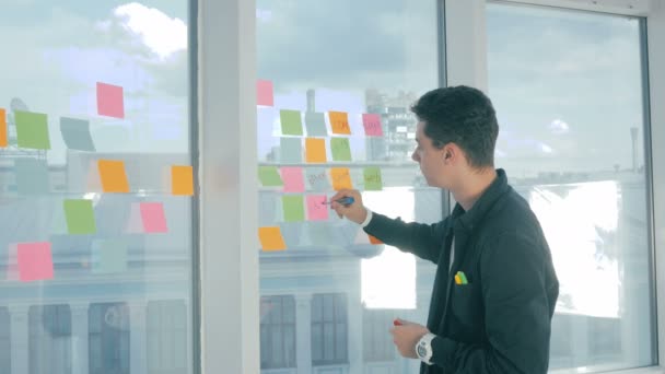 Junger gutaussehender Mann, der auf klebrige Zettel am Fenster eines modernen Büros schreibt. Brainstorming, Benennung. — Stockvideo