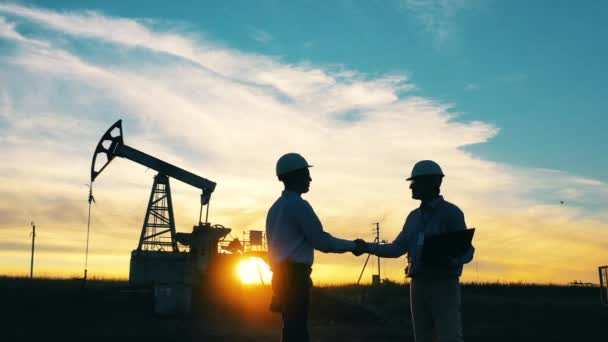 Two engineers shaking hands in front of oil pumpjack at sunset background — Stock Video