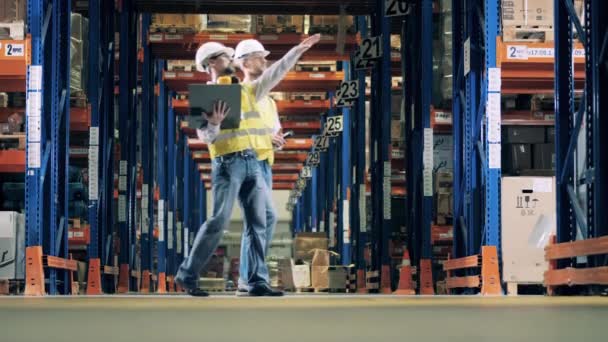 Двое рабочих с ноутбуком пересекают складской зал — стоковое видео