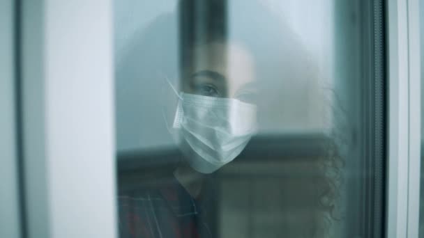 Νεαρή γυναίκα με ιατρική μάσκα κοιτάζει από το παράθυρο. — Αρχείο Βίντεο
