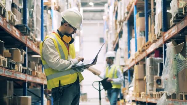 Storeman in uniform typing on laptop among warehouse shelves — Stock Video