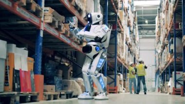 Depo robotu arka planda iki işçiyle birlikte karton kutularda barkodları tarıyor.