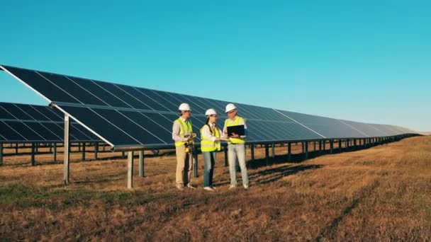 Снимок дрона с участием трех специалистов по солнечной энергии, обсуждаемых на солнечной электростанции — стоковое видео