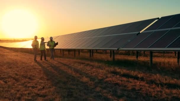 Três especialistas em energia solar caminhando por um parque solar ao pôr-do-sol. Drone disparado — Vídeo de Stock