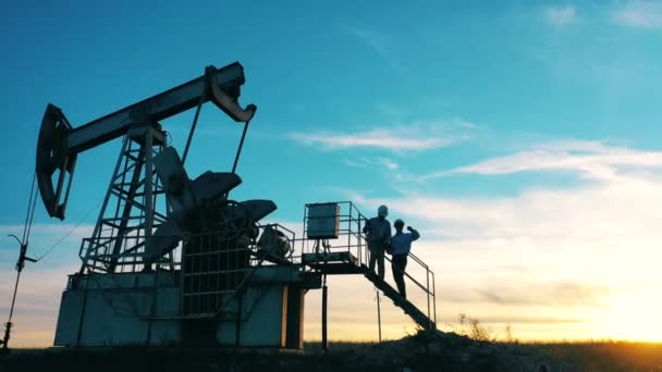 Dos trabajadores petroleros inspeccionando un campo petrolero al atardecer — Vídeo de stock