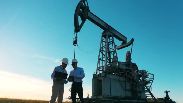 Dos trabajadores de la industria petrolera discutiendo en un campo petrolero con una bomba de petróleo — Vídeos de Stock