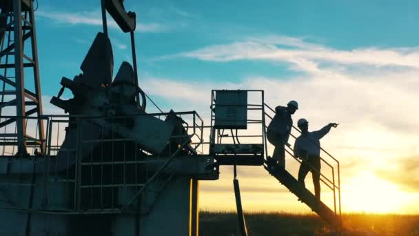 Zwei Ölarbeiter unterhalten sich bei Sonnenuntergang an einer Ölpumpe — Stockvideo
