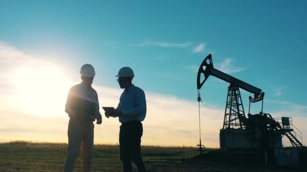 Dos petroleros hablando en un campo petrolífero al atardecer — Vídeos de Stock