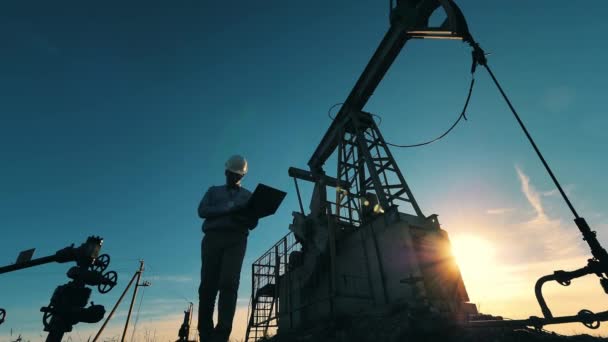 Εργάτης της πετρελαϊκής βιομηχανίας περπατά μπροστά από ένα γεωτρύπανο στο φως του ήλιου. Αργή κίνηση — Αρχείο Βίντεο