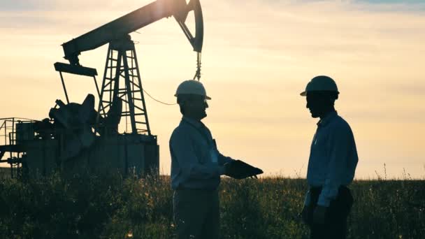 Two oil workers shaking hands with oil pumpjack operating in background — Video