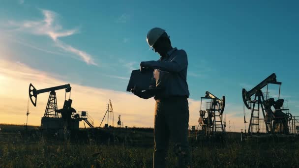 Trabajador del petróleo escribiendo en el ordenador portátil entre las zapatillas de aceite en el campo petrolífero — Vídeo de stock