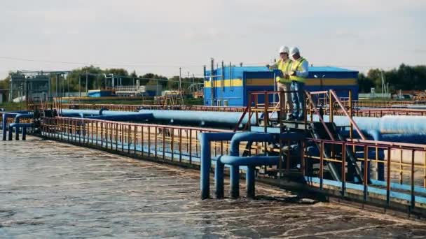 Dos especialistas hablando mientras están de pie en un puente sobre el depósito de limpieza de aguas residuales — Vídeos de Stock