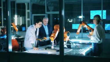 Üçüncüsü kontrol cihazını kullanarak robot operasyonlarını yönetirken iki mühendis konuşuyor.