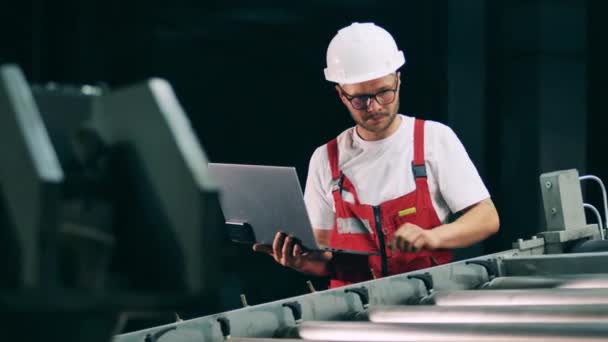 Trabajador de metalurgia con un portátil está observando un transportador de trabajo — Vídeos de Stock